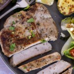 A plate with a partially sliced grilled pork chop garnished with herbs, accompanied by grilled zucchini, a lemon wedge, and a side salad with tomatoes, cucumbers, and lettuce.