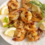 A plate of grilled shrimp garnished with lemon wedges and fresh parsley on a light-colored surface.