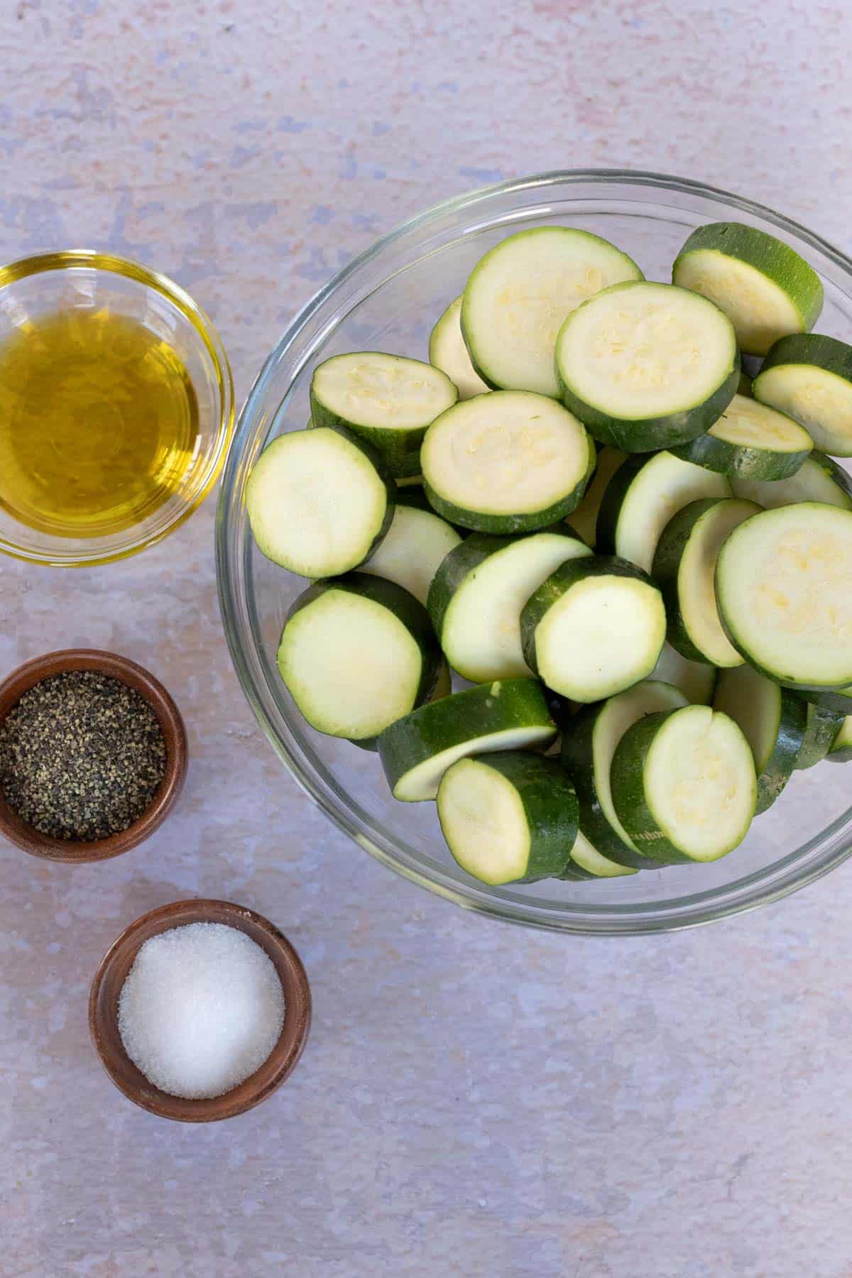 A bowl of sliced zucchini with a glass bowl of olive oil, and small wooden bowls of black pepper and salt on a light-colored surface.