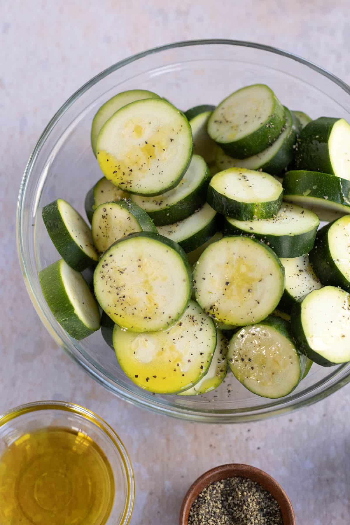 A clear bowl filled with sliced zucchini seasoned with olive oil and black pepper. Nearby, there is a small bowl of olive oil and a small bowl of ground black pepper.