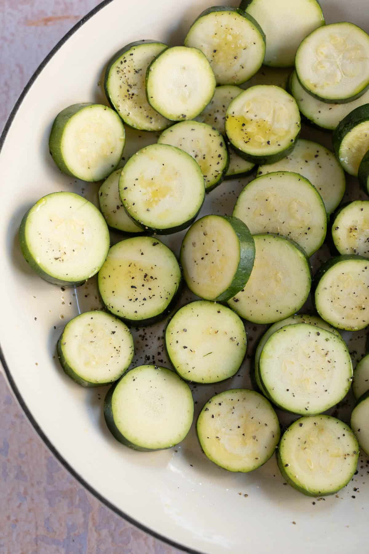 A white bowl filled with sliced zucchini seasoned with oil, salt and pepper.