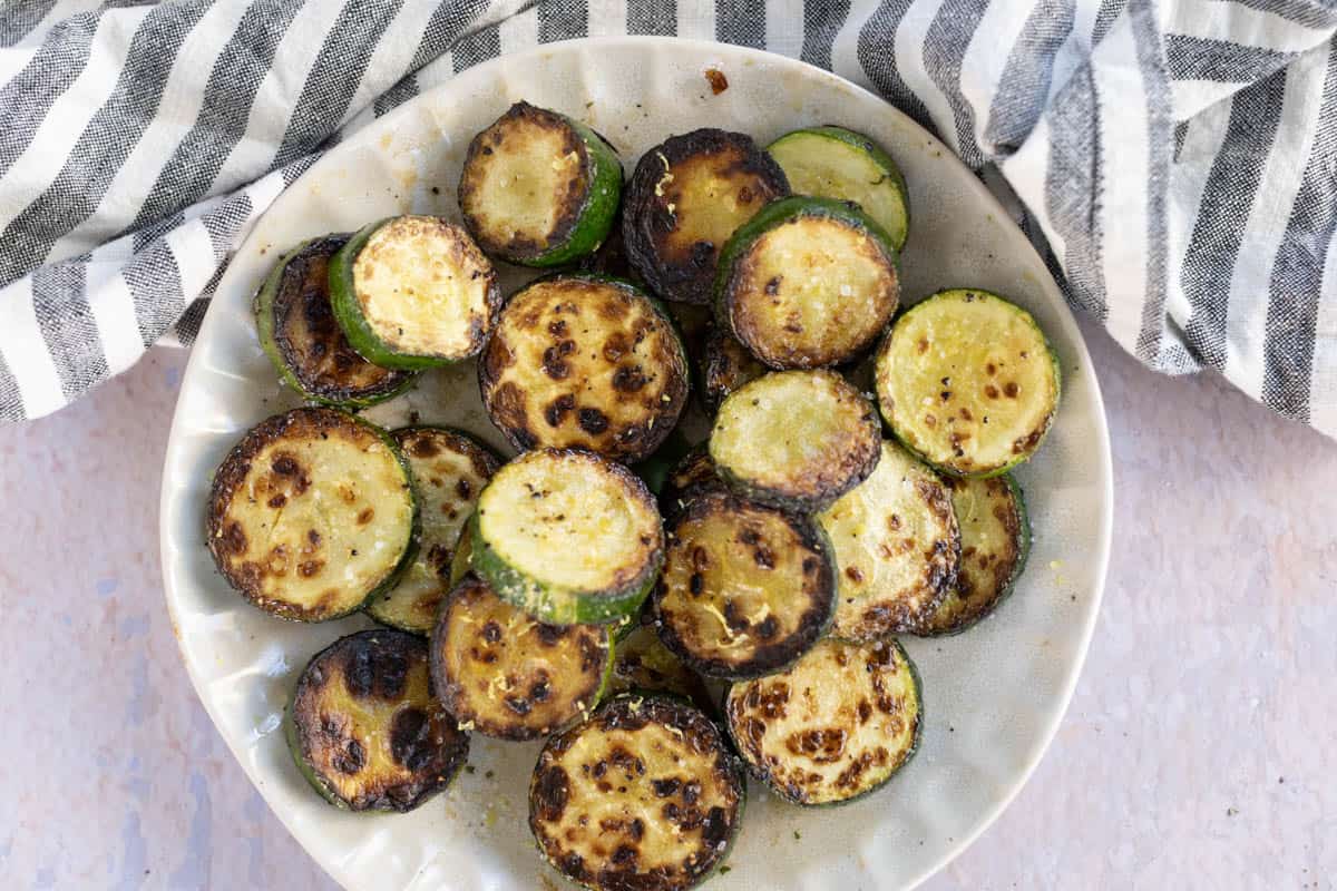 A plate of saute&eacute;d zucchini slices with charred edges, placed on a light-colored surface next to a black and white striped cloth.
