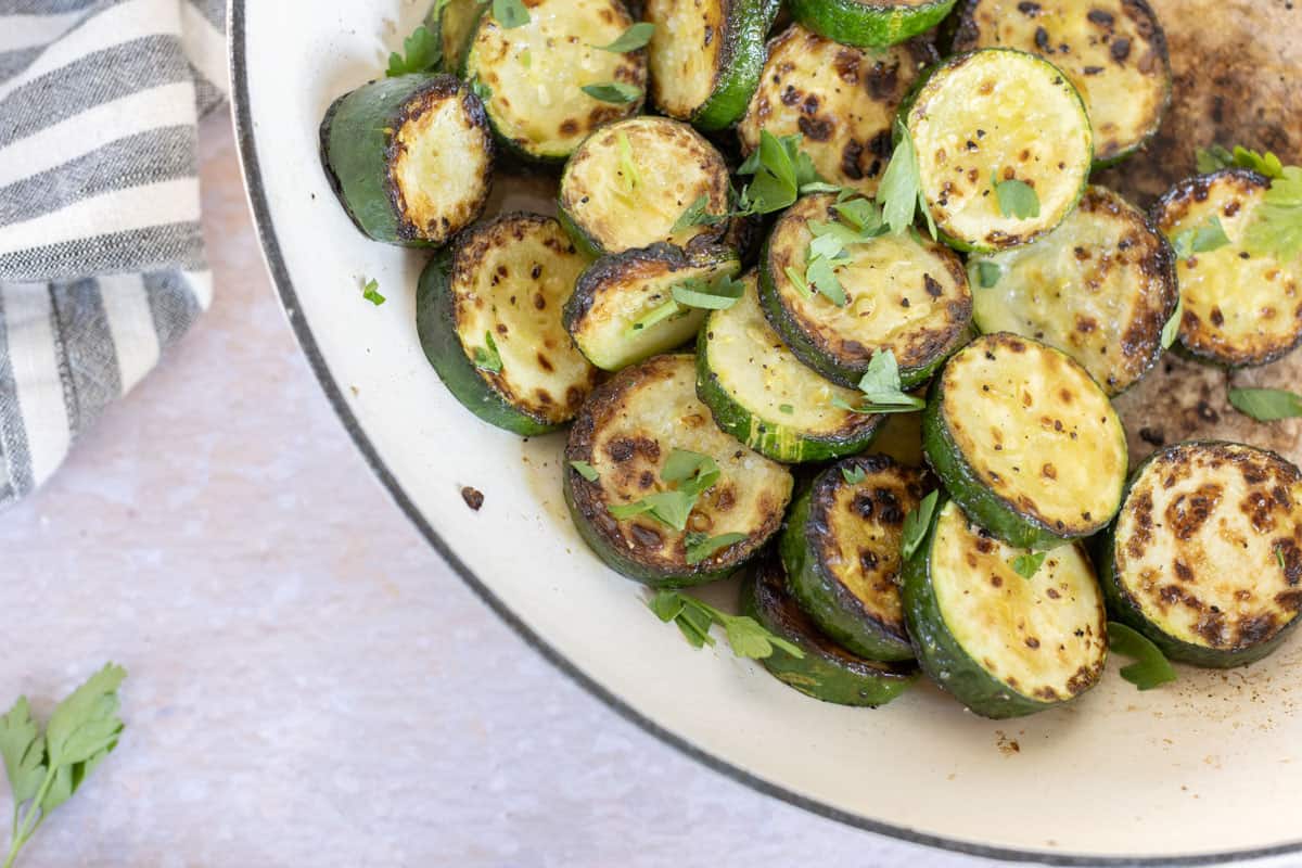 A white bowl filled with roasted zucchini slices garnished with chopped parsley is placed on a light-colored surface next to a striped cloth.