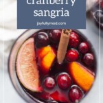 A glass of spiced cranberry sangria garnished with fresh cranberries, orange slices, and a cinnamon stick. Perfect for pairing with grilled chicken thighs. The label at the top reads "spiced cranberry sangria" from joyfullymad.com.