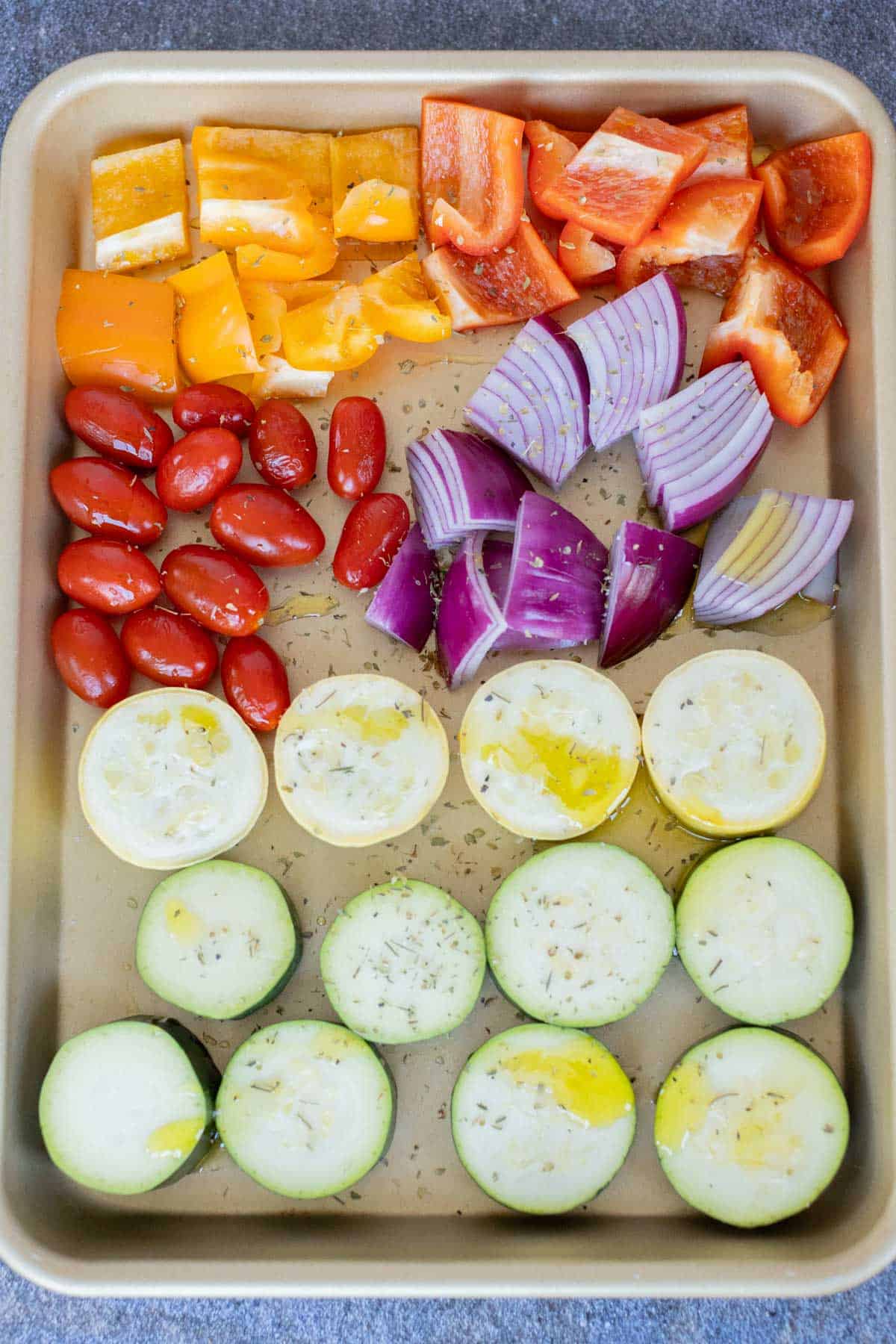 A baking sheet with assorted raw vegetables: chopped bell peppers, grape tomatoes, red onion wedges, and sliced zucchini, drizzled with oil and herbs, ready for roasting.