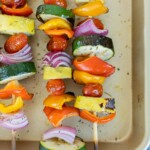 Four grilled vegetable skewers with cherry tomatoes, zucchini, yellow squash, red onion, and bell peppers are arranged on a beige baking sheet.