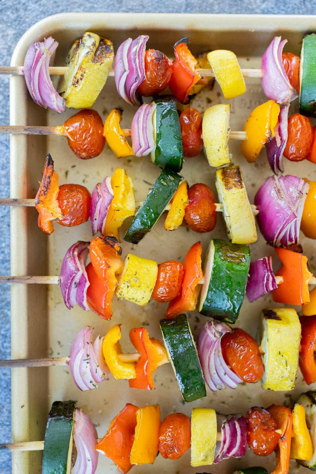 A baking tray with six colorful, grilled vegetable skewers, including zucchini, red onion, bell peppers, and cherry tomatoes. The skewers are arranged in neat rows ready to be served.