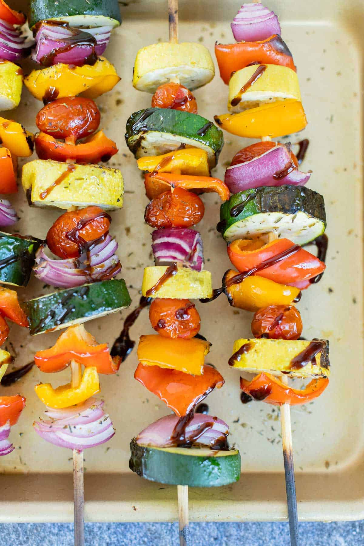 Colorful, grilled vegetable skewers with zucchini, bell peppers, cherry tomatoes, and red onions, drizzled with a balsamic glaze, arranged on a baking sheet.