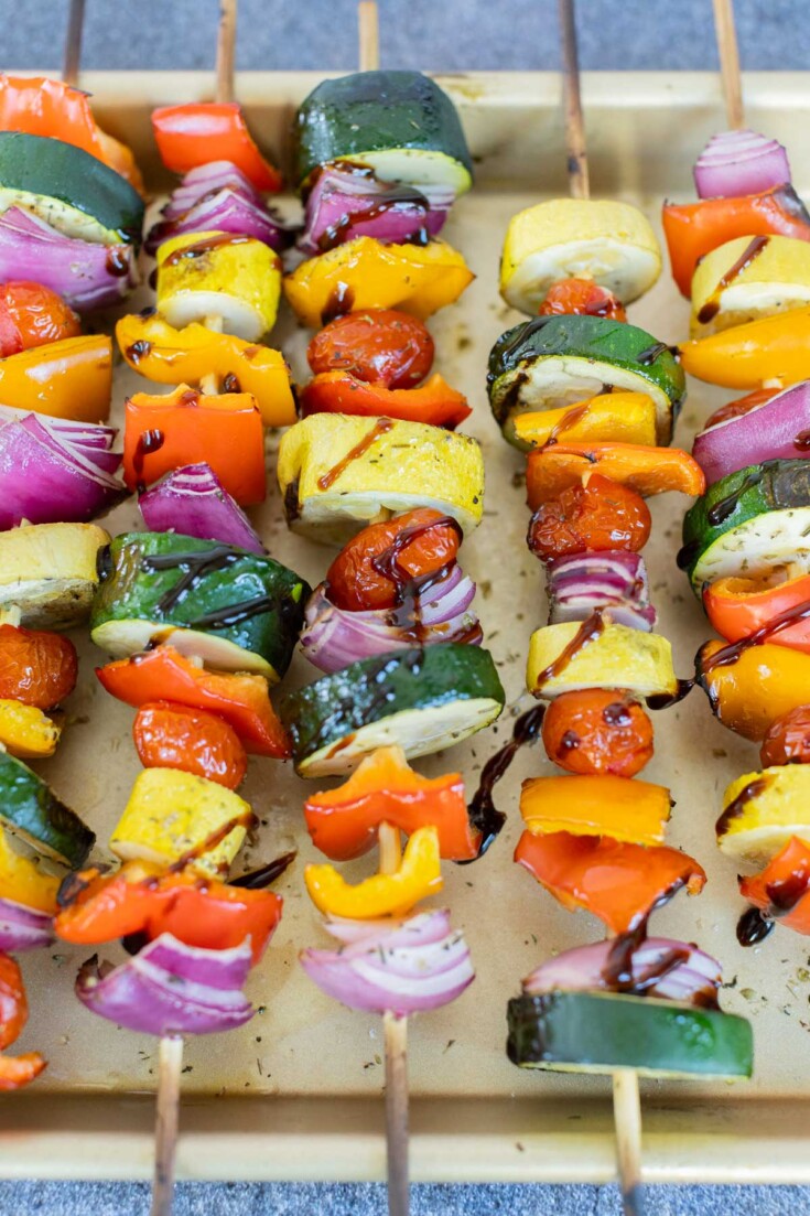 A tray of grilled vegetable skewers with zucchini, bell peppers, cherry tomatoes, and red onions, drizzled with balsamic glaze.