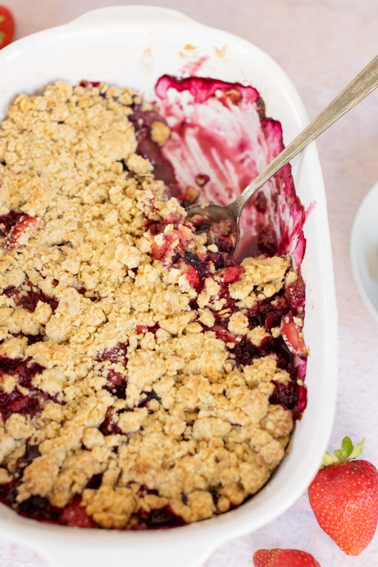 A baked mixed-berry crisp in a white rectangular dish, topped with a golden crumb layer and a spoon placed inside. Some strawberries are visible next to the dish.