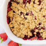 A close-up of a mixed-berry crisp in a white baking dish with a topping of golden brown oat crisp. Two whole strawberries are placed beside the dish.
