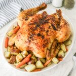 A roasted chicken garnished with herbs is served on a white plate. Surrounding the chicken are roasted potatoes and baby carrots. A striped cloth, salt, pepper, and a serving spoon are in the background.