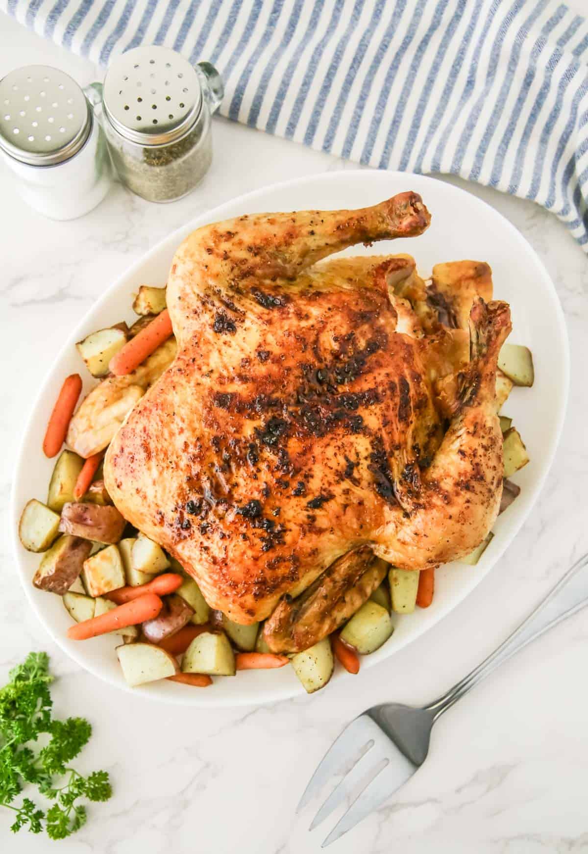 A roasted chicken is served on a white platter with carrots and potatoes. Salt and pepper shakers along with a striped cloth are in the background, and a metal fork is placed beside the platter.