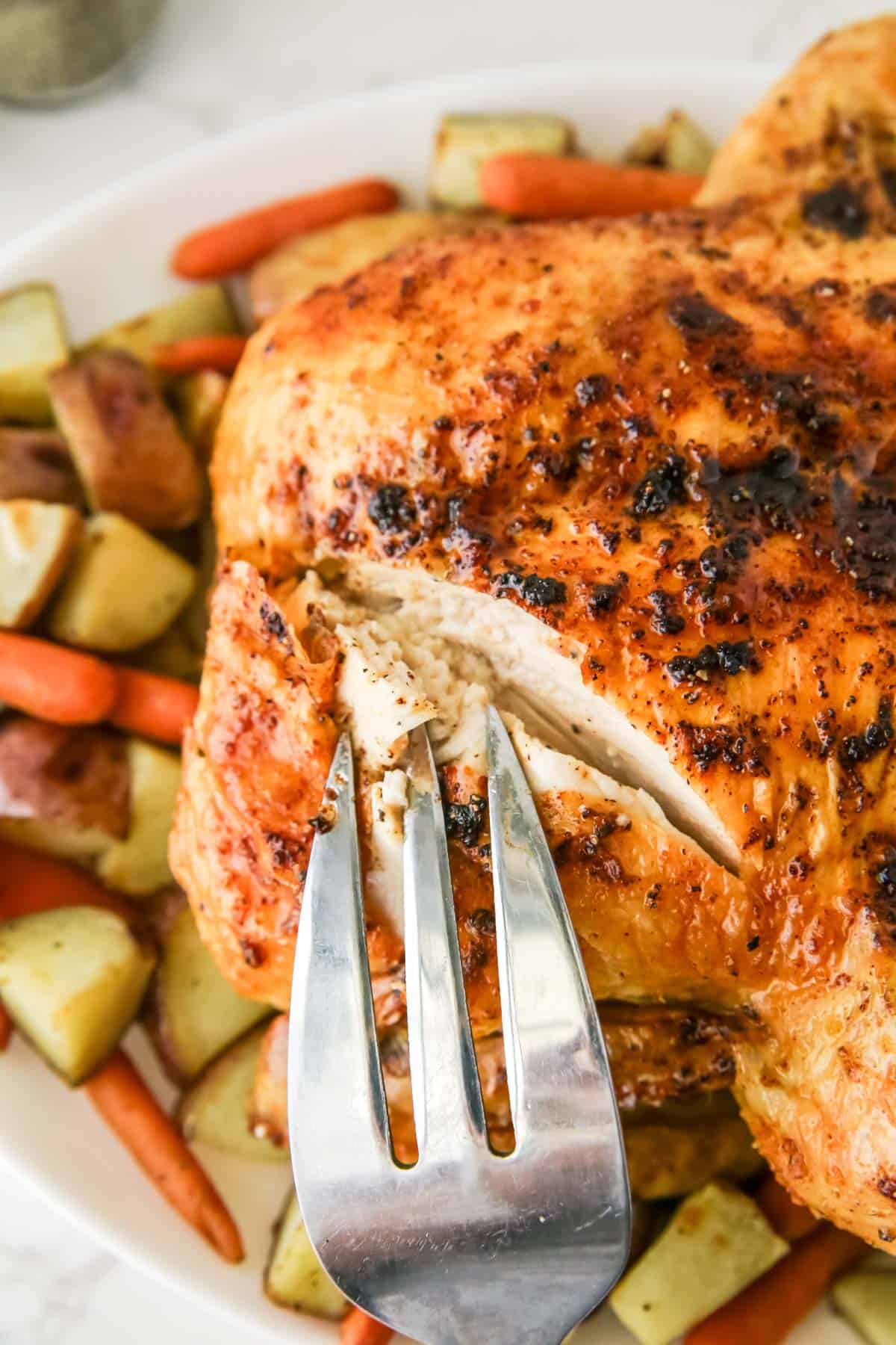 A roasted chicken, garnished with herbs and spices, is being cut with a fork. The dish is surrounded by roasted potatoes and carrots.