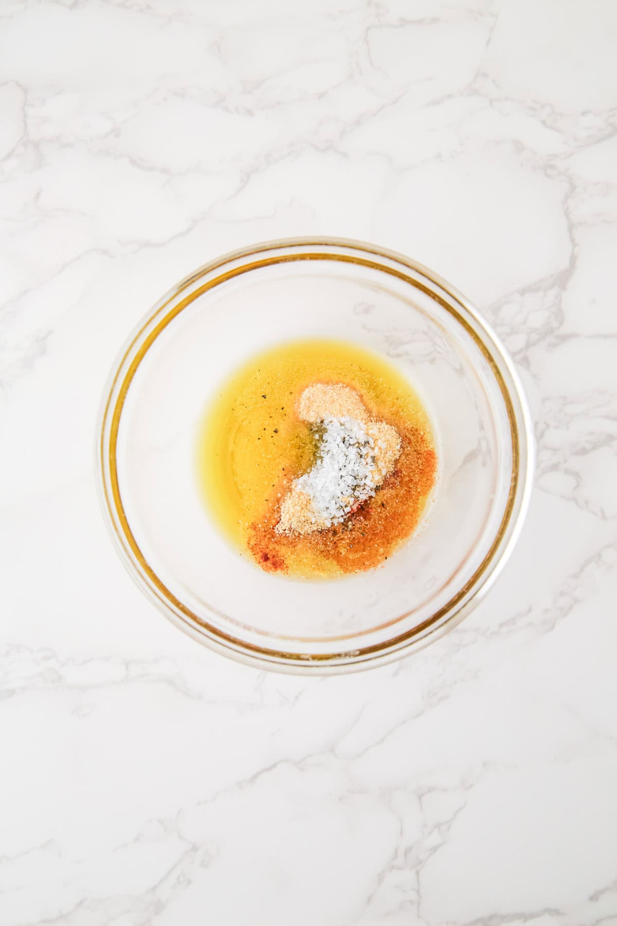 A clear glass bowl containing a mix of oil, spices, and salt on a white marble surface.