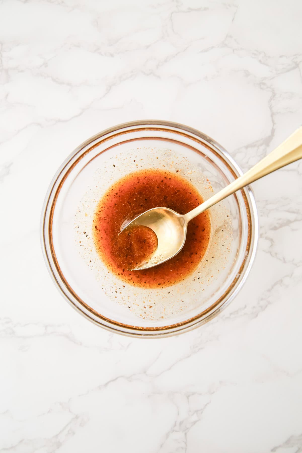 A glass bowl containing orange-brown liquid with seasoning, mixed with a silver spoon, placed on a marble surface.
