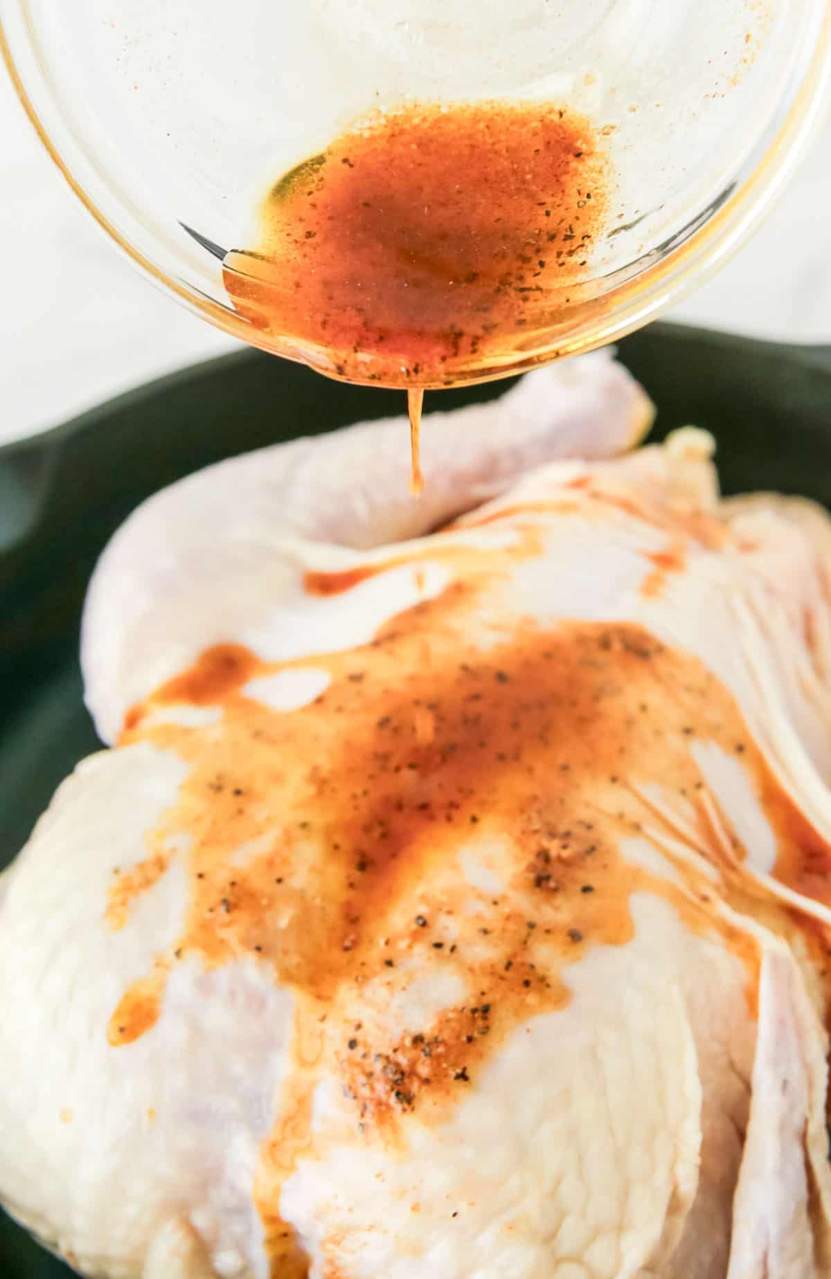 Close-up of a raw chicken being marinated with a brown, seasoned liquid being poured from a glass bowl above.