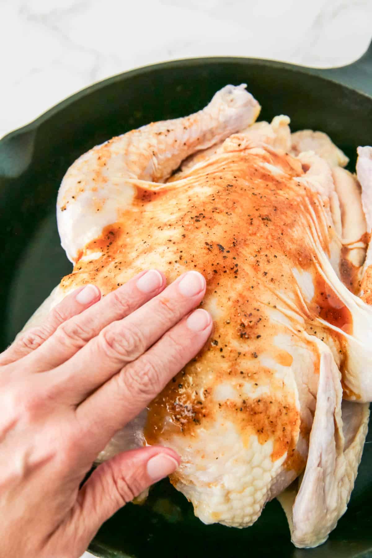 A hand is pressing down on a raw, seasoned whole chicken in a dark-colored pan.