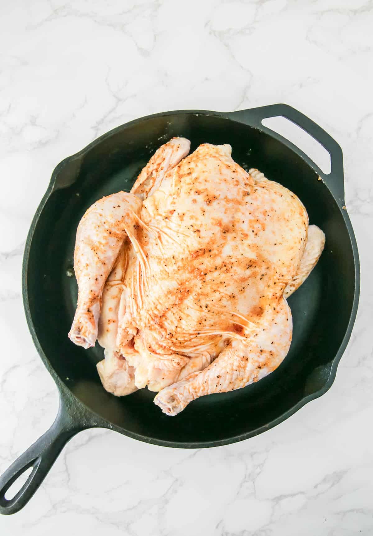 A raw, seasoned chicken placed in a black cast iron skillet on a marble countertop.
