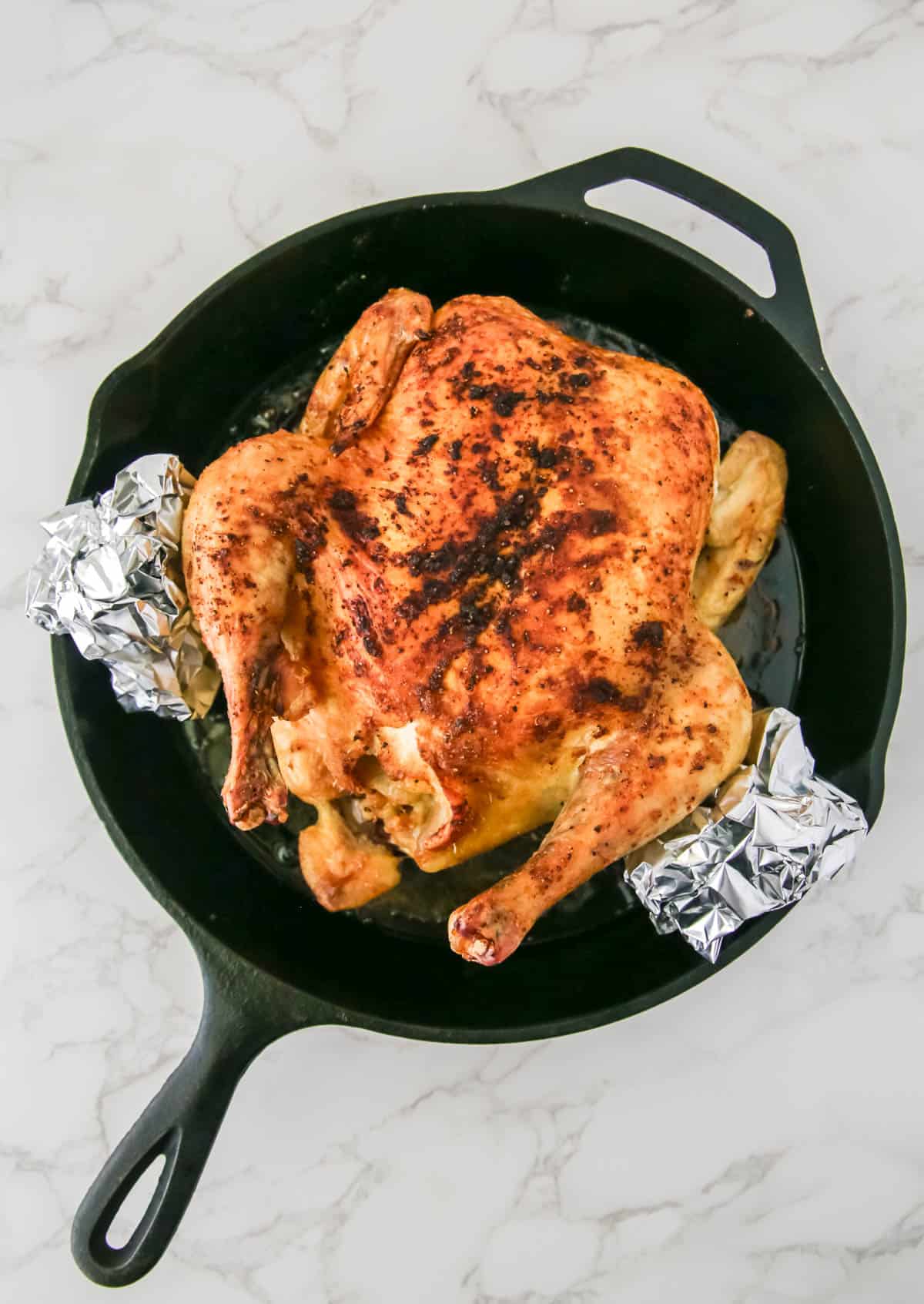 A whole roasted chicken in a cast-iron skillet, with its wing tips wrapped in aluminum foil, placed on a marble surface.