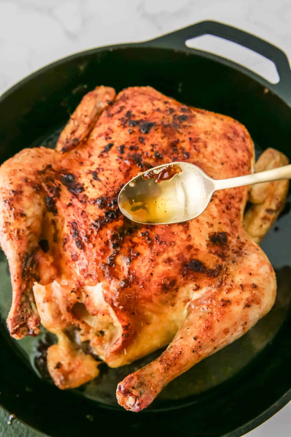 A roasted whole chicken in a black skillet with seasoning visible on the surface. A spoon with liquid sauce is being drizzled over the chicken.