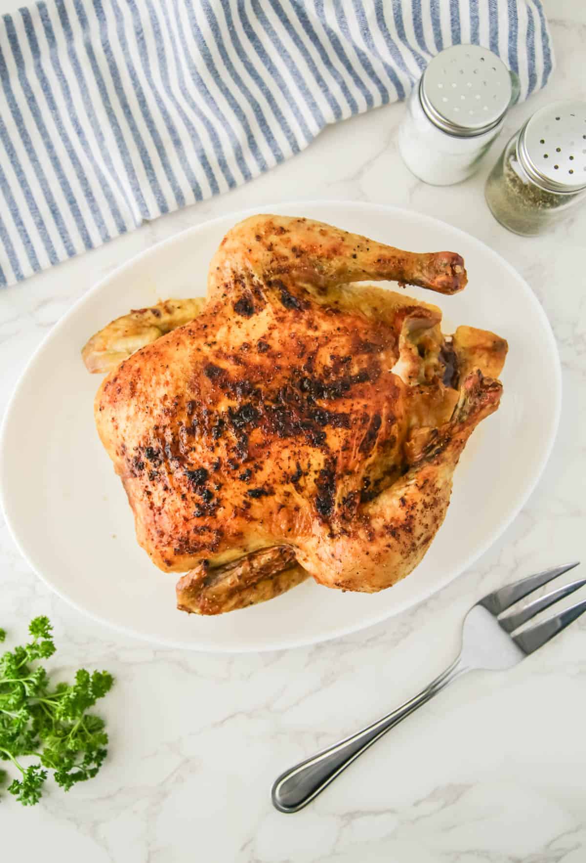 A roasted chicken on a white plate, surrounded by a napkin, salt and pepper shakers, parsley, and a fork on a marble surface.