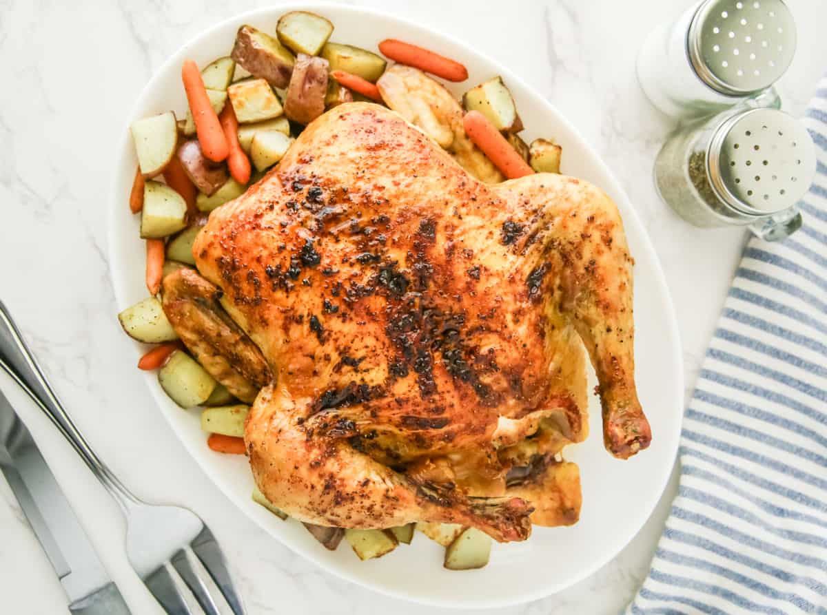 A roasted chicken surrounded by roasted potatoes and carrots on a white plate. A fork and knife are next to the plate along with salt and pepper shakers and a blue-striped cloth.