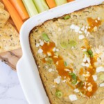 A dish of baked buffalo chicken dip topped with crumbled cheese and green onions, served with carrot and celery sticks, and tortilla chips on a marble surface.