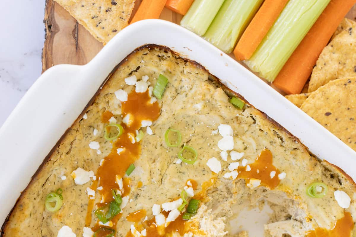 A close-up of a baked buffalo chicken dip in a white dish, topped with chopped green onions and crumbled cheese. Nearby are carrot sticks, celery sticks, and tortilla chips.