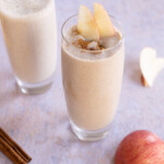 Two glasses of caramel apple smoothies; one topped with apple slices and cinnamon, an apple and cinnamon sticks are in the foreground.