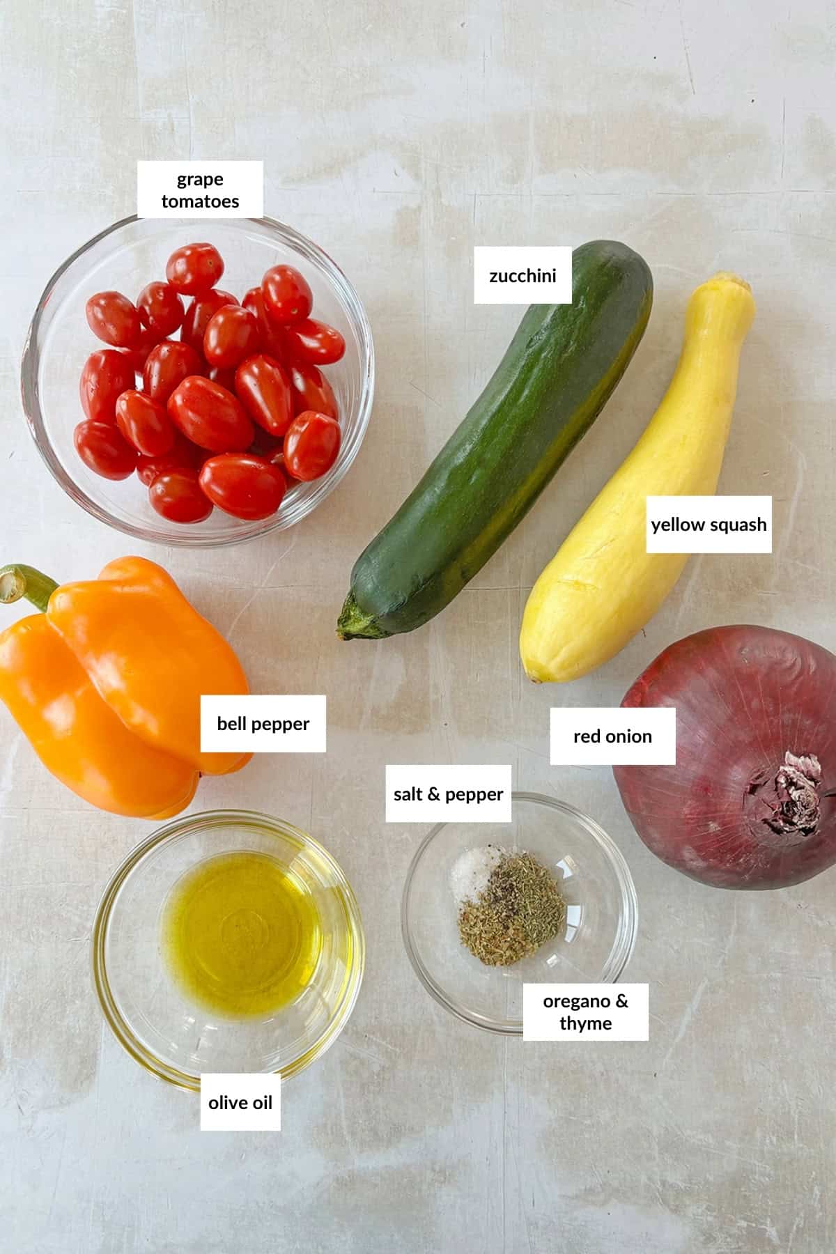 Assorted vegetables and seasonings including grape tomatoes, zucchini, yellow squash, bell pepper, red onion, olive oil, salt & pepper, oregano & thyme, arranged on a light surface.