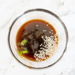 A glass bowl containing a dark sauce with sesame seeds and chopped green onions on a white marble surface.