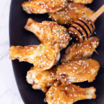 A black platter holds several glazed chicken wings sprinkled with sesame seeds. A honey dipper is placed beside the wings.