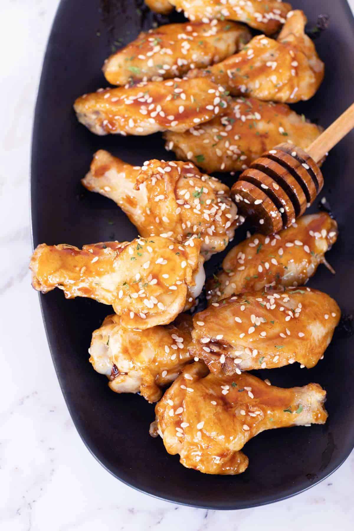 A black platter holds several glazed chicken wings sprinkled with sesame seeds. A honey dipper is placed beside the wings.