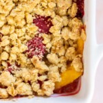 Close-up of a fruit crisp in a white baking dish, showing a golden-brown, crumbly topping with visible pieces of peaches and raspberries underneath.