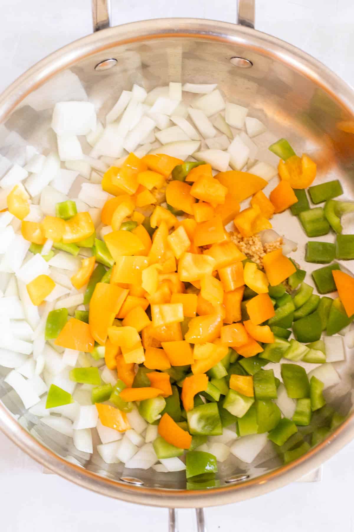 A stainless steel pan holds chopped onions, yellow and green bell peppers, and minced garlic ready for cooking.