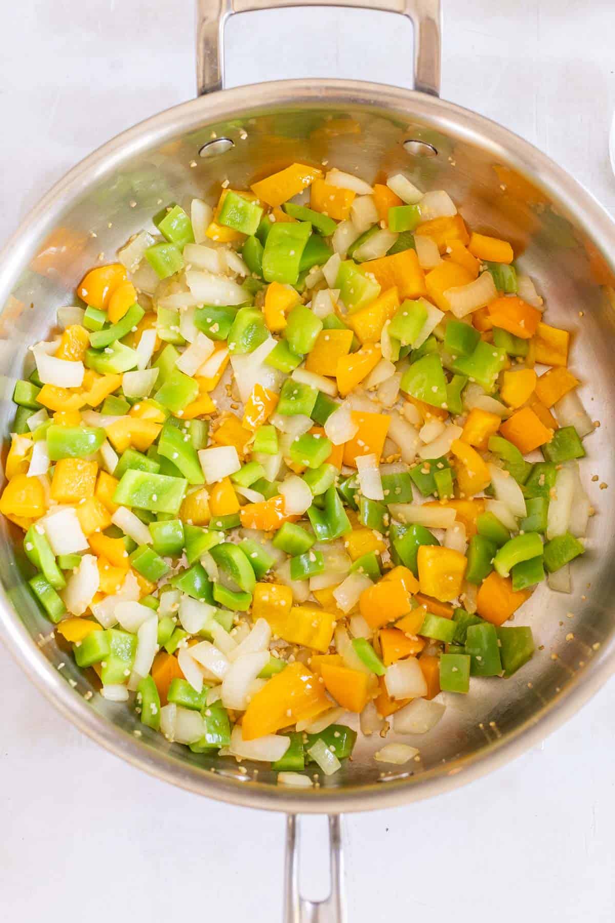 Chopped green and yellow bell peppers, onions, and garlic cooking in a large stainless steel pan.