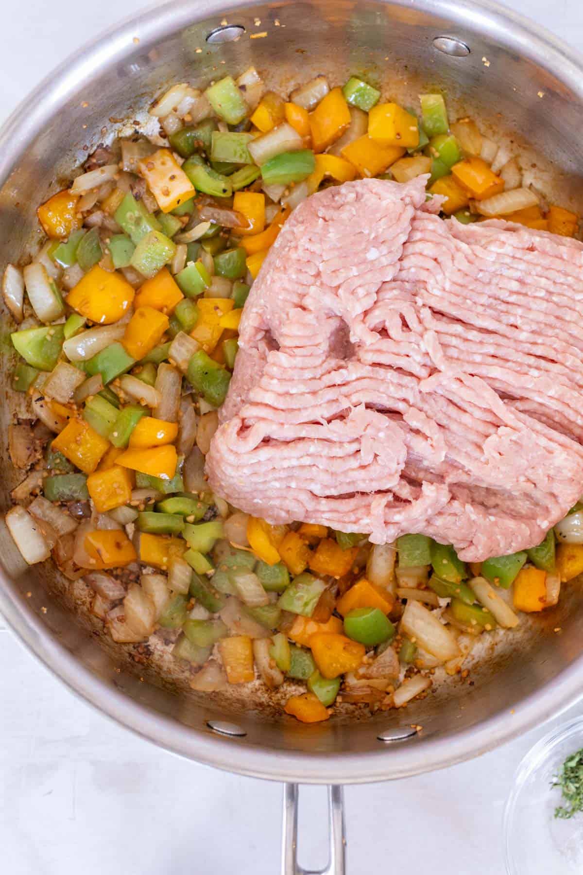 A skillet with chopped onions, green and yellow bell peppers, and a block of raw ground turkey on top.