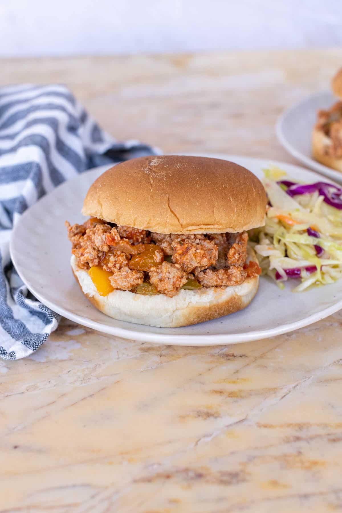 A turkey sloppy joe sandwich with a side of coleslaw is on a white plate, placed on a marble countertop. A striped cloth is partially visible in the background.