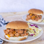 Two plates of turkey sloppy joe burgers with coleslaw on a marble surface. A striped napkin is seen on the left side of the image.