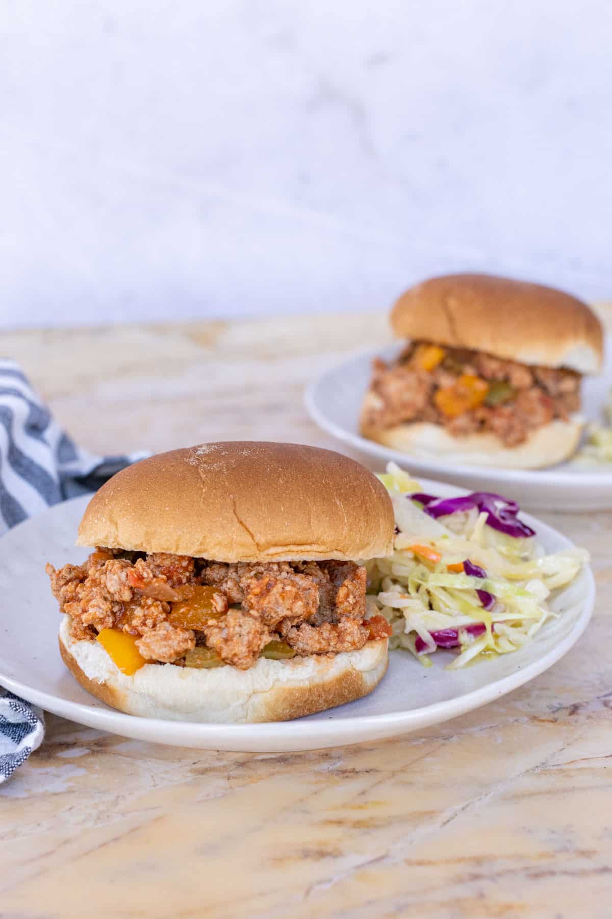 Two plates of turkey sloppy joe burgers with coleslaw on a marble surface. A striped napkin is seen on the left side of the image.