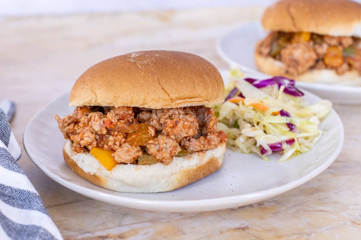 A sloppy joe sandwich with minced meat and vegetables sits on a white plate next to a serving of coleslaw. A second sandwich on another plate is partially visible in the background.