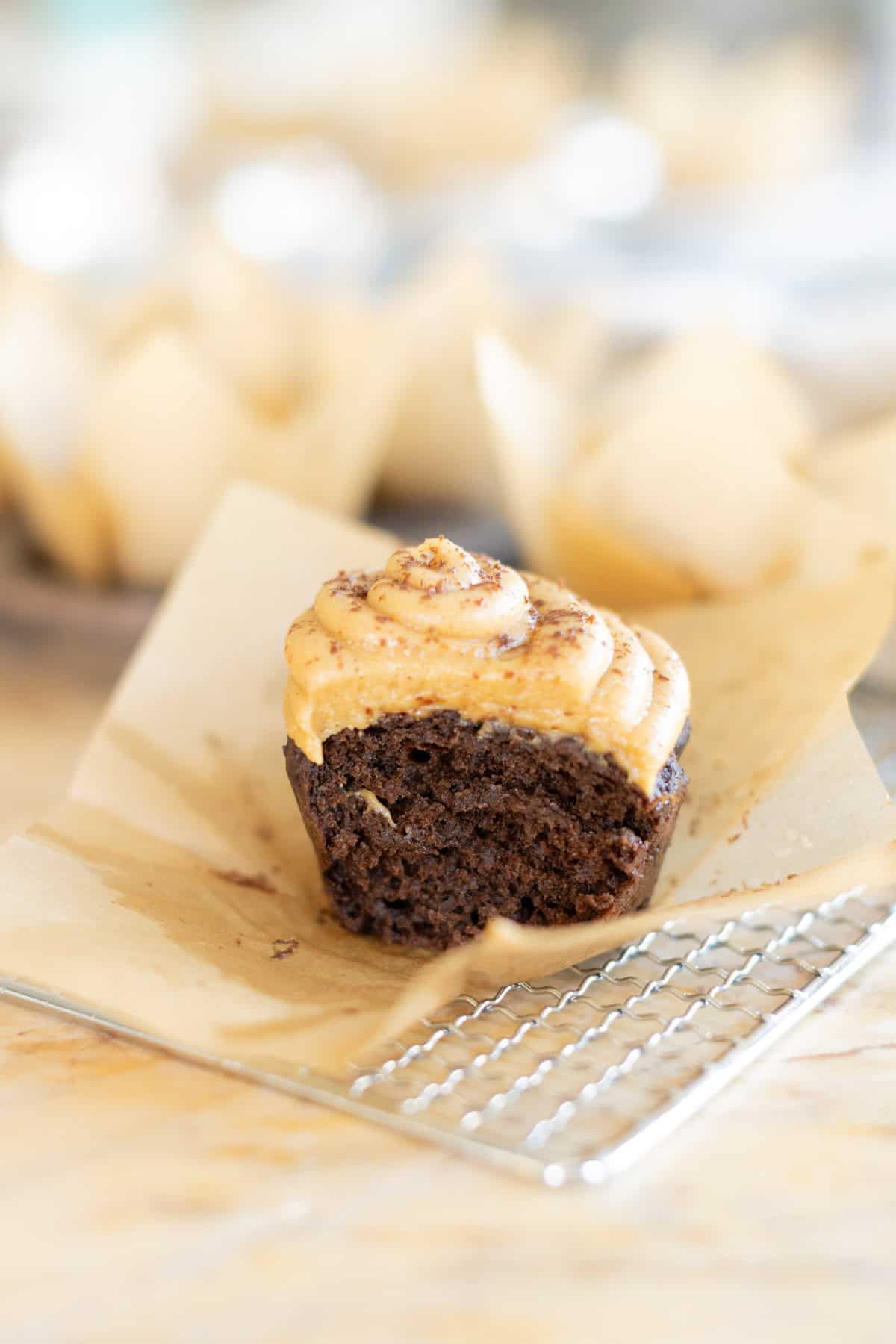 A chocolate cupcake with peanut butter frosting, with a bite taken from it, on parchment paper and a metal cooling rack.
