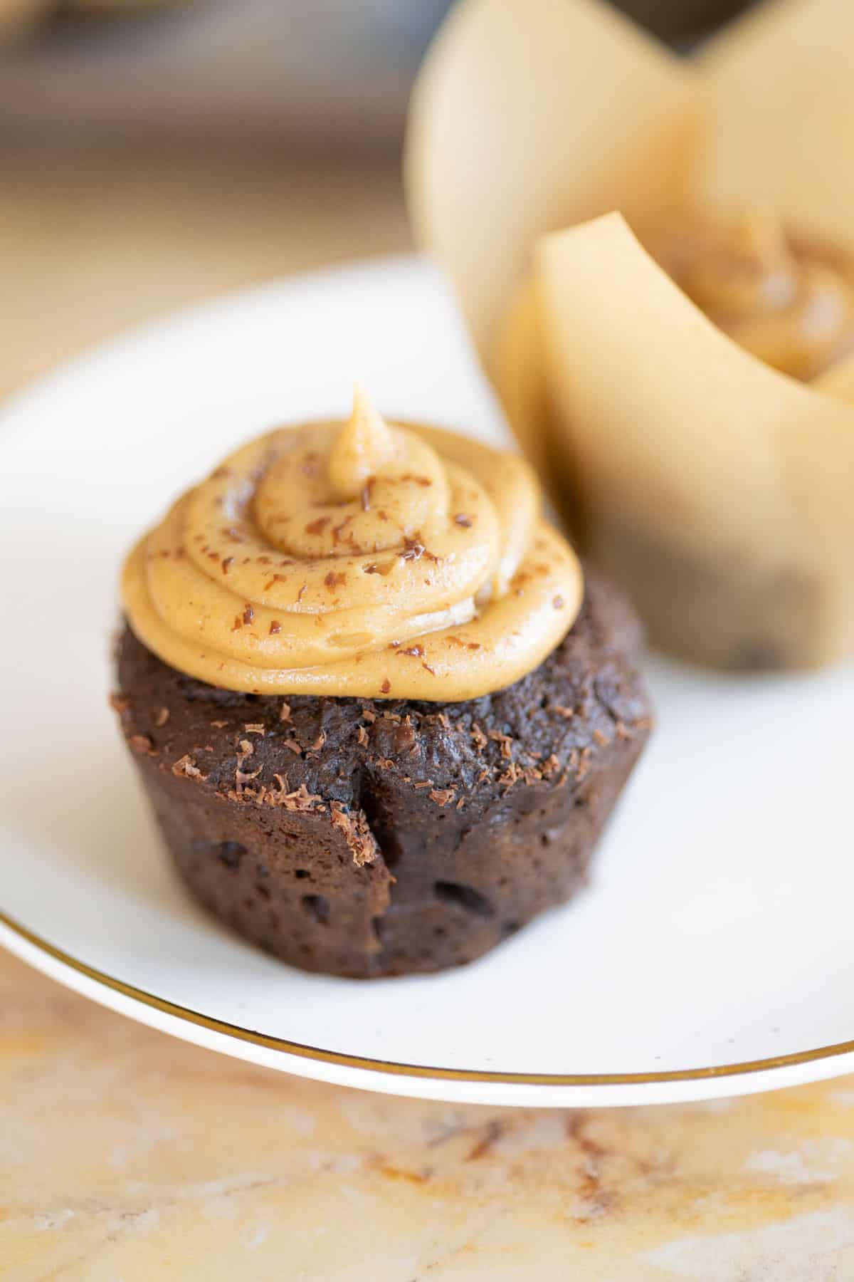 A chocolate muffin topped with a swirl of peanut butter frosting on a white plate, with another muffin partially wrapped in parchment paper in the background.