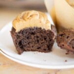 A chocolate muffin with a bite taken out of it, topped with peanut butter frosting, is placed on a white plate. Another partially visible muffin is in the background.