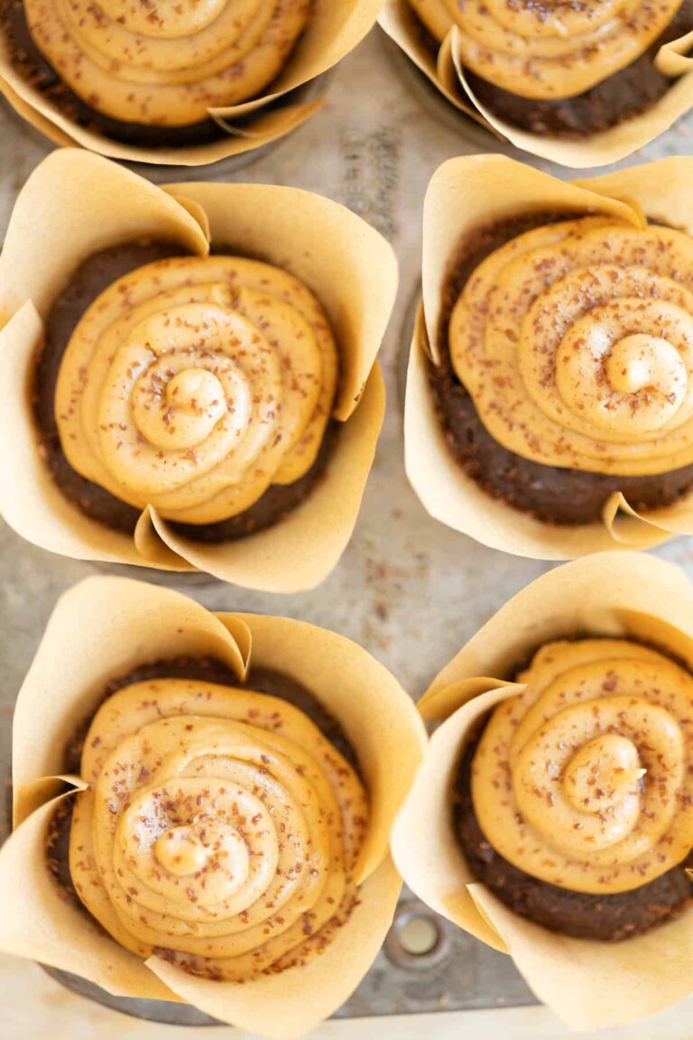 Close-up view of six chocolate muffins in paper cups, topped with swirls of peanut butter frosting and cocoa powder, displayed in a metal muffin tray.