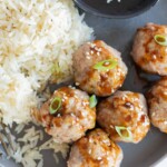 A plate with white rice, five honey sriracha meatballs garnished with sauce, sesame seeds, and green onions, and a small black bowl of dipping sauce.