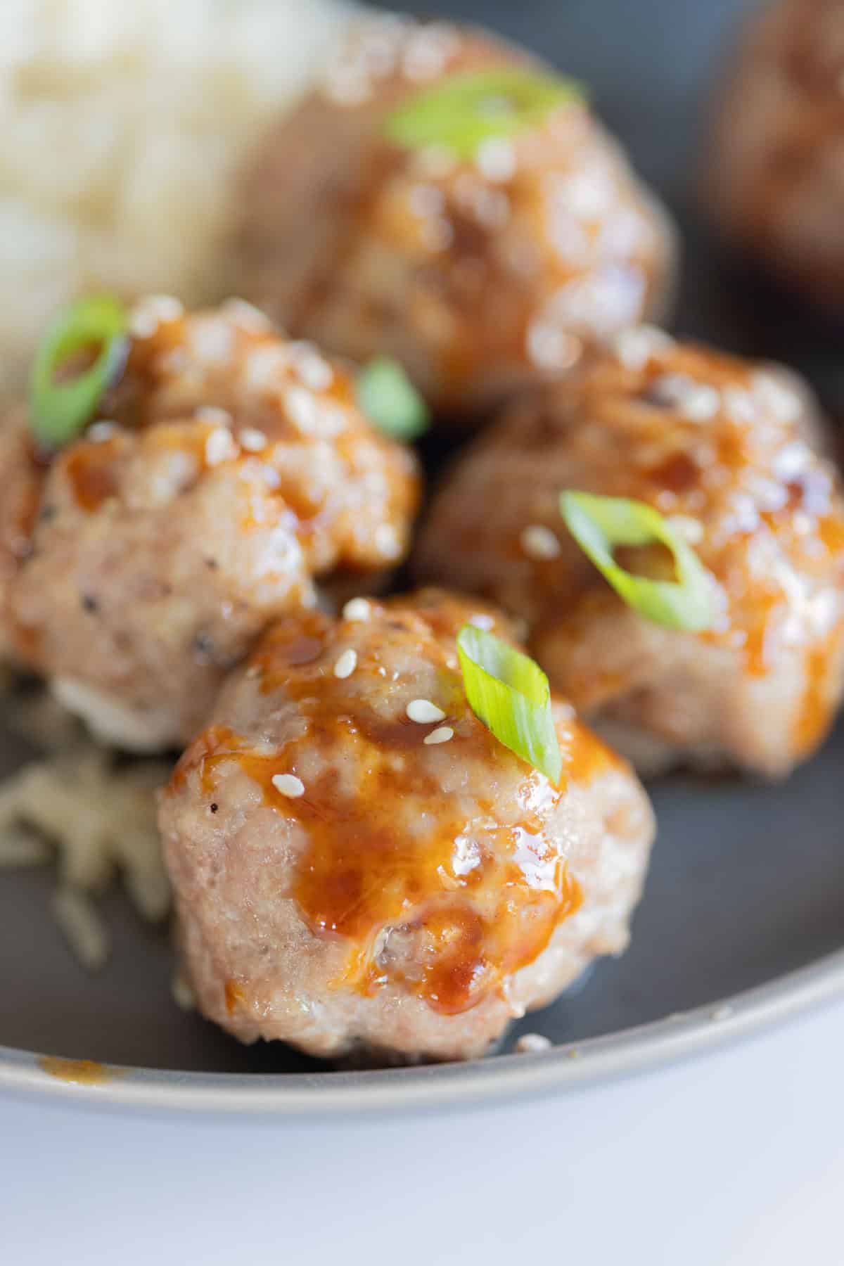 Close-up of four meatballs topped with sauce, sesame seeds, and green onions on a gray plate, with a side of rice in the background.