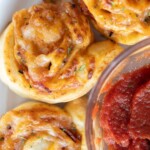 Close-up of freshly baked pepperoni pinwheels with melted cheese and herbs, next to a glass bowl of marinara sauce.