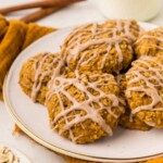 A plate of pumpkin oatmeal cookies drizzled with icing, set on a white surface with a yellow cloth, cinnamon sticks, and a small bowl of oats in the background.
